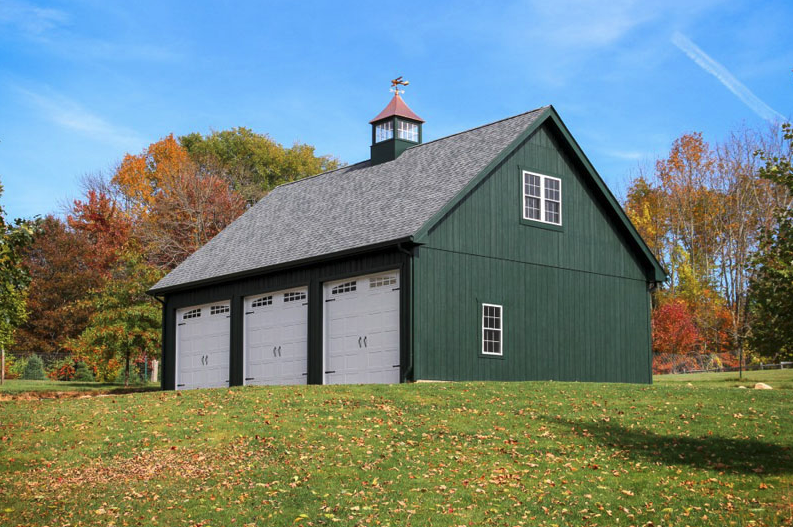A green barn with three garage doors is in the middle of a grassy field.