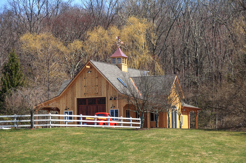 A large wooden barn is surrounded by trees and a white fence.