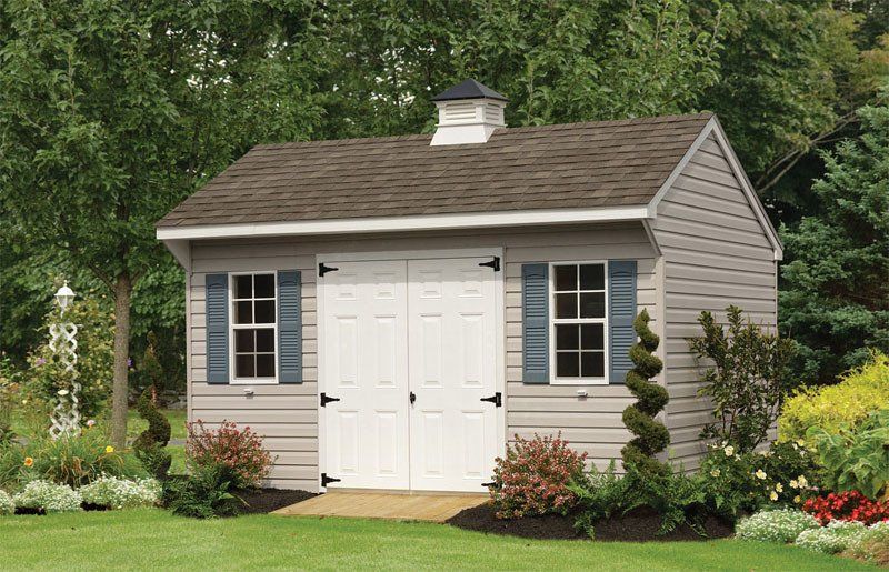 A small shed with blue shutters is sitting in the middle of a lush green yard.