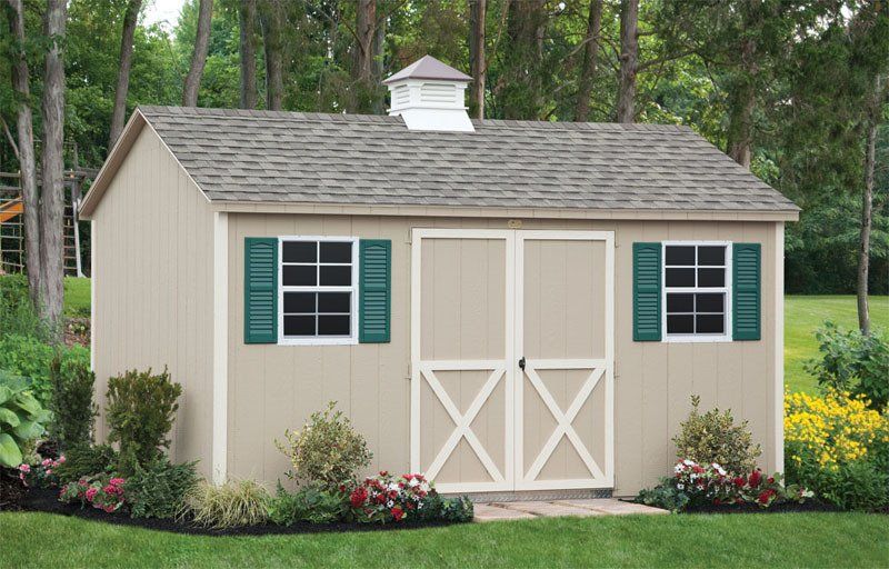 A beige shed with green shutters is sitting in the middle of a lush green yard.