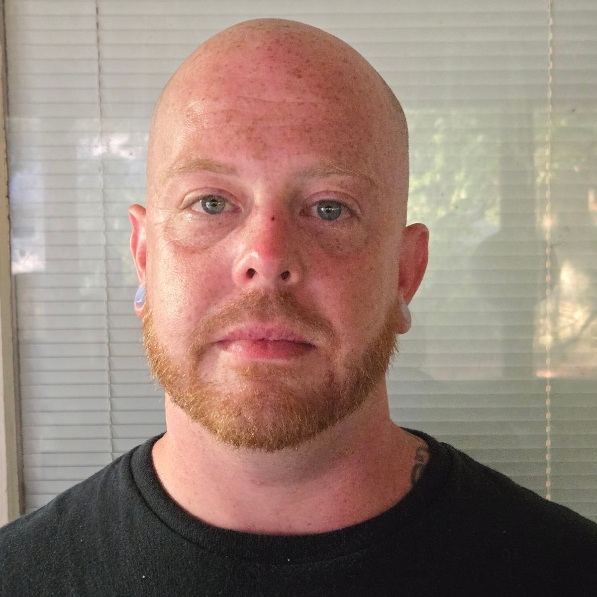 A bald man with a beard is wearing a black shirt and looking at the camera.