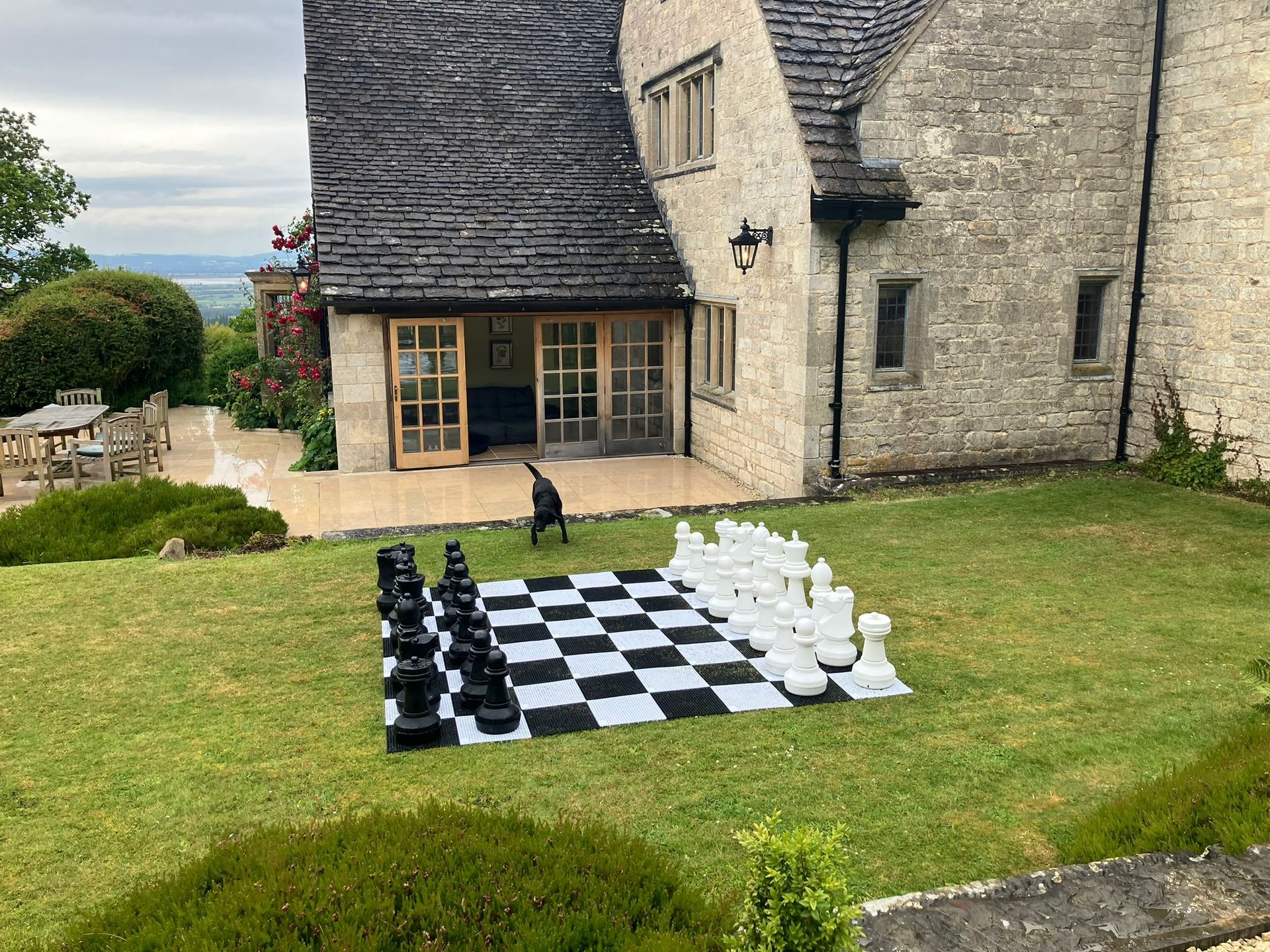 White and black chess board on a garden of grass.  With giant chess pieces.  A large brick home in the back ground.