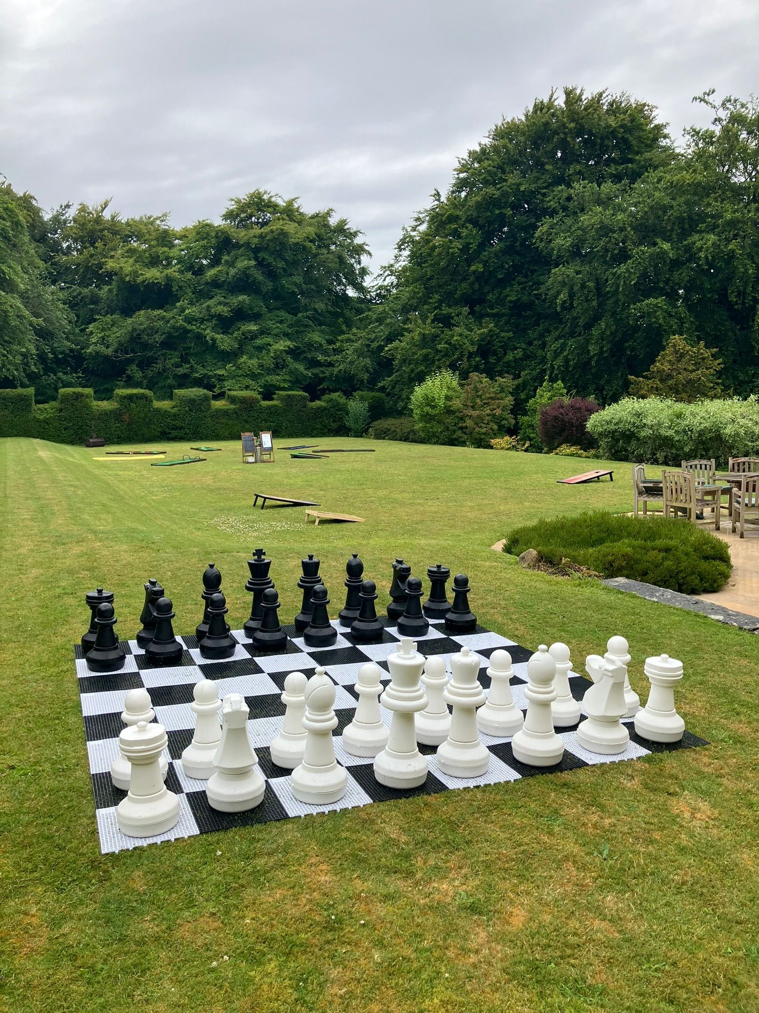 White and black chess board on garden of grass.  With giant chess pieces.  crazy golf and garden furniture in the back ground