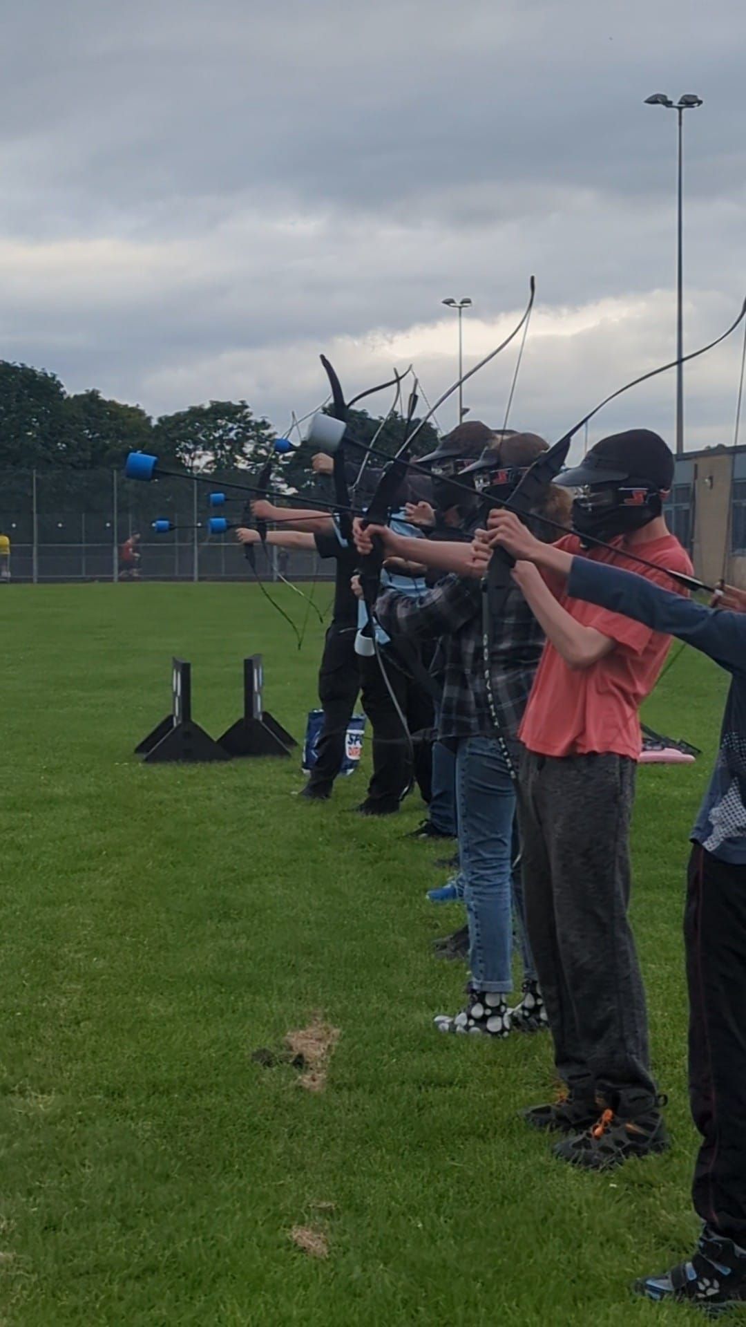 A group of people in a line in safety head gear, holding archery tag bows and arrows, waiting to shoot.