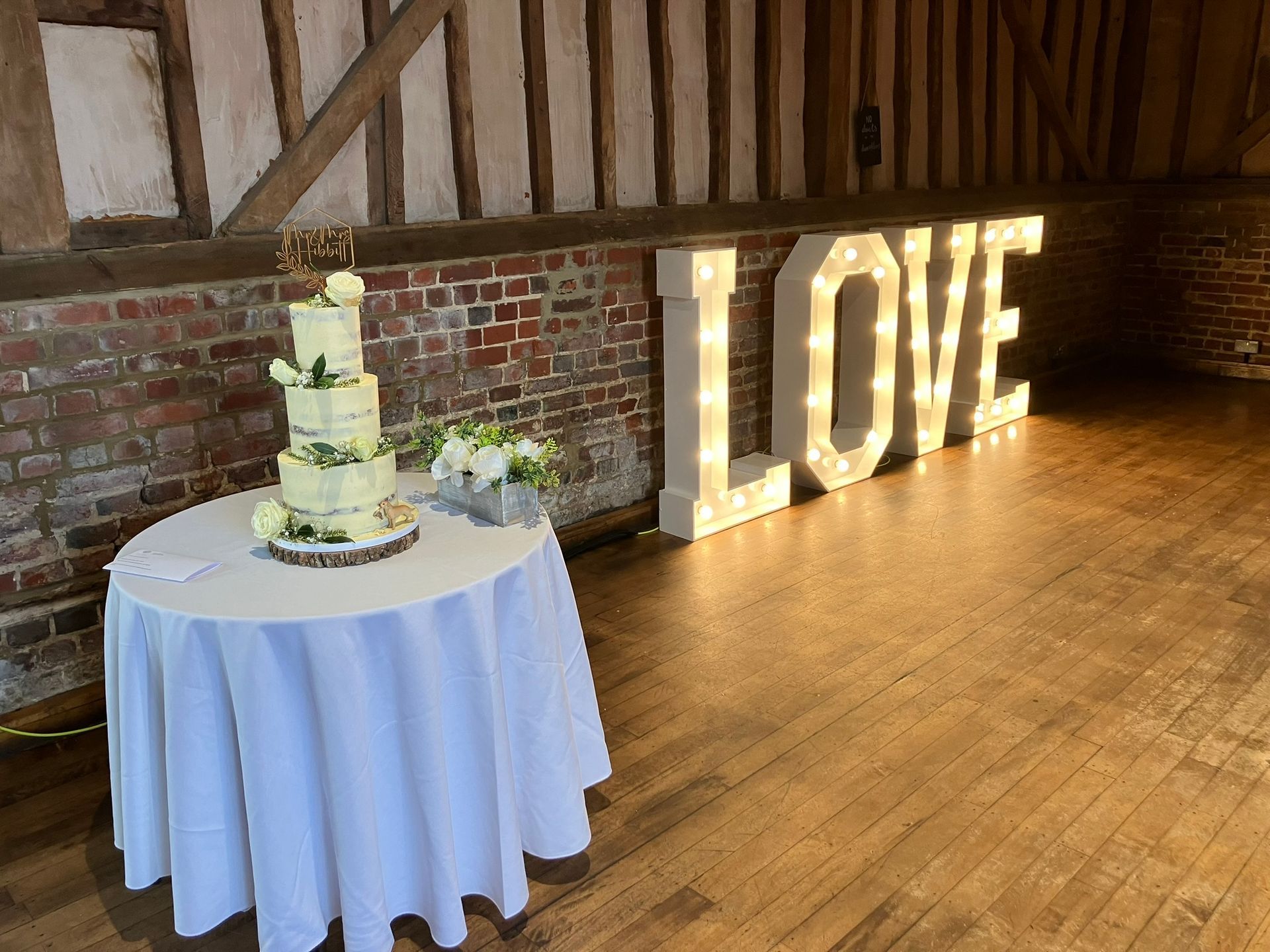LOVE lights in a barn on a dance floor next to a table with  a wedding cake on it.