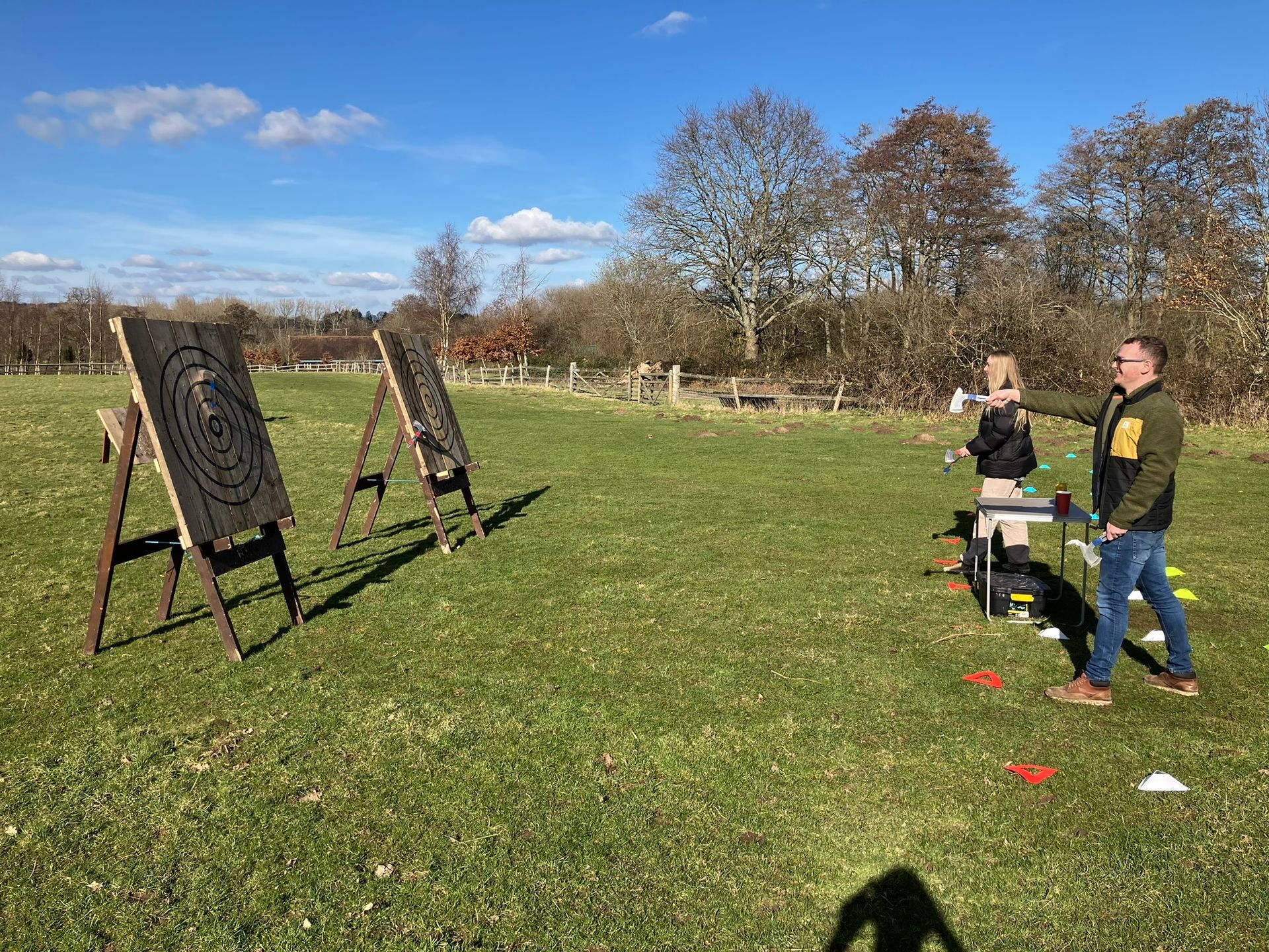 2 wooden axe throwing target on a field with a man and a woman throwing an axe towards it