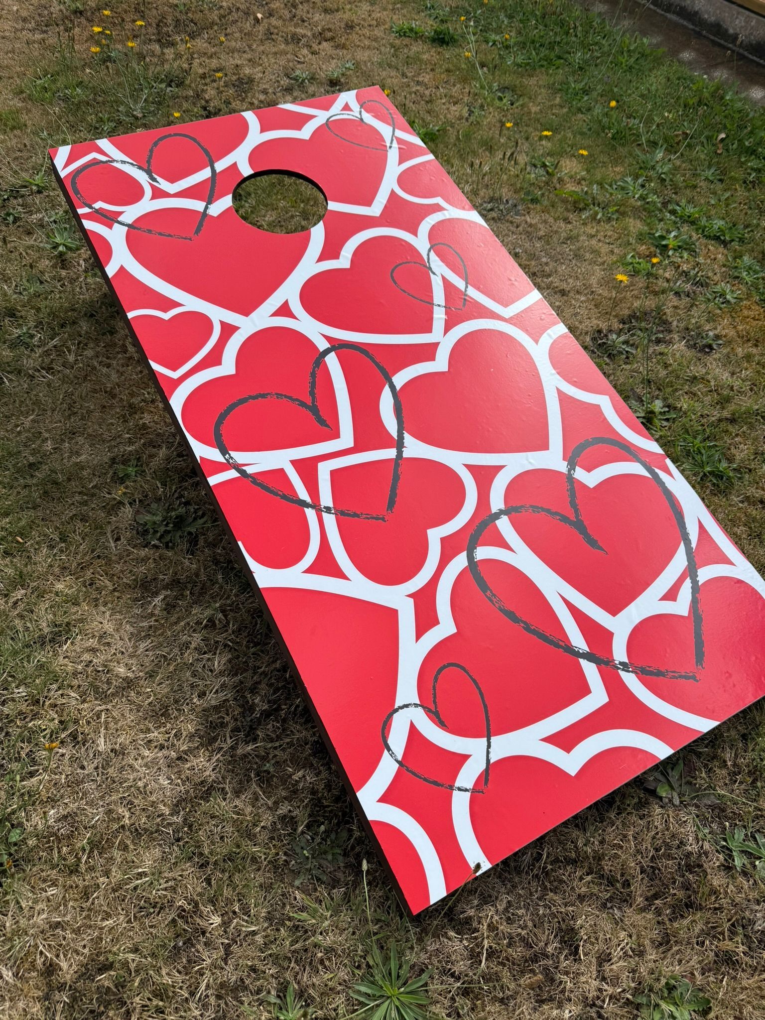 A red hearts themed cornhole board on grass