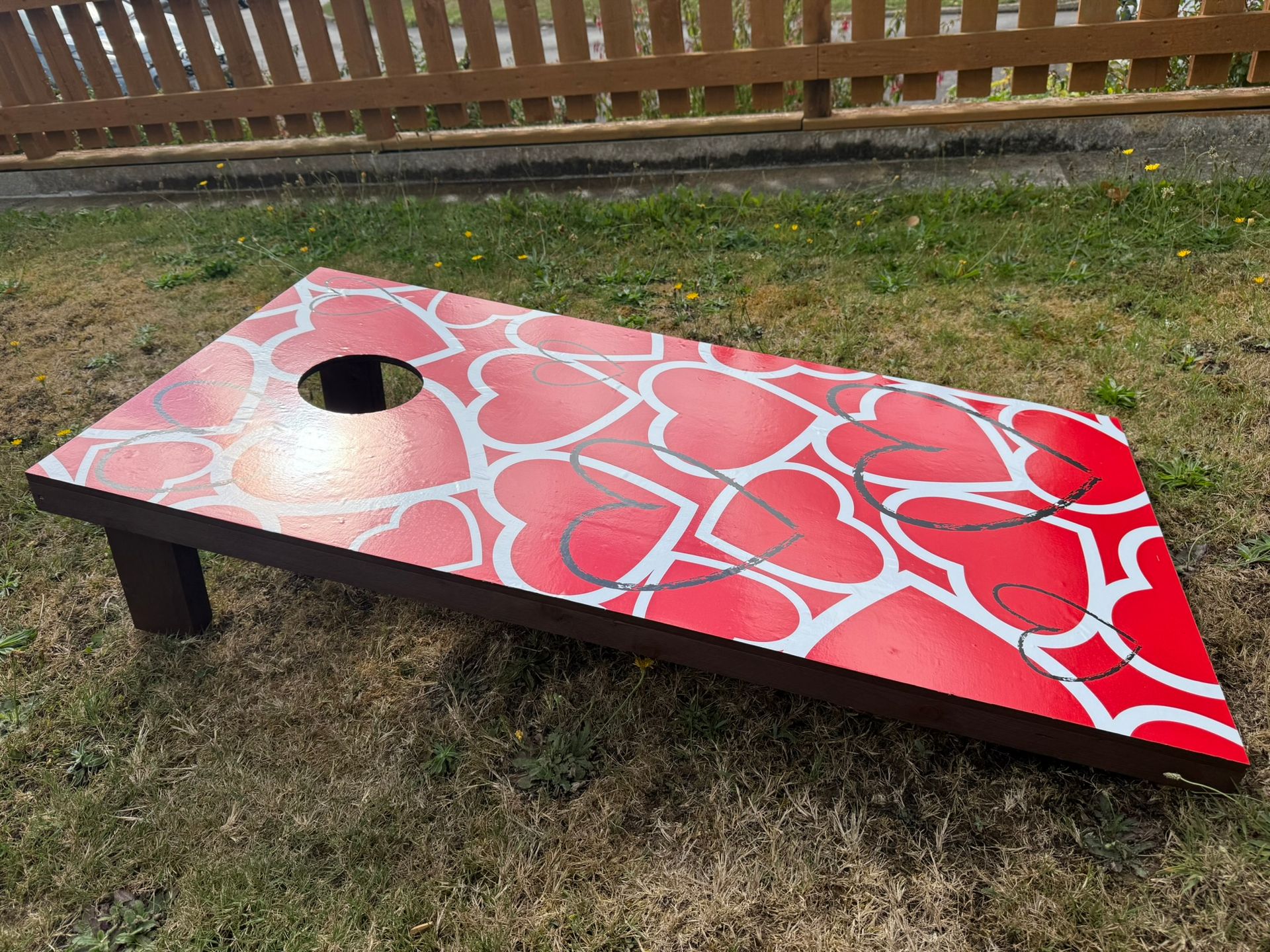 A red hearts themed cornhole board on grass