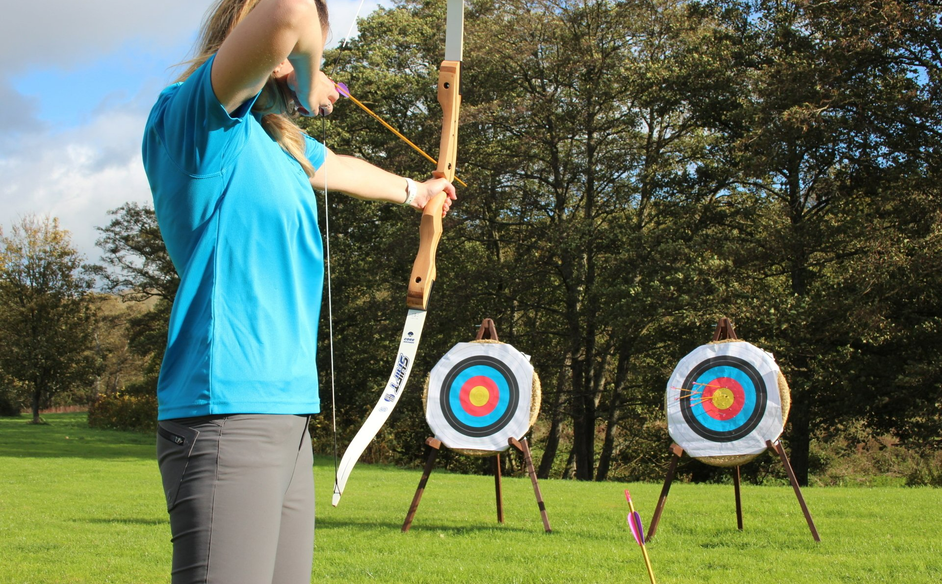 A young woman holding a bow, aimed at an archery target.  There are 2 targets in the photo.