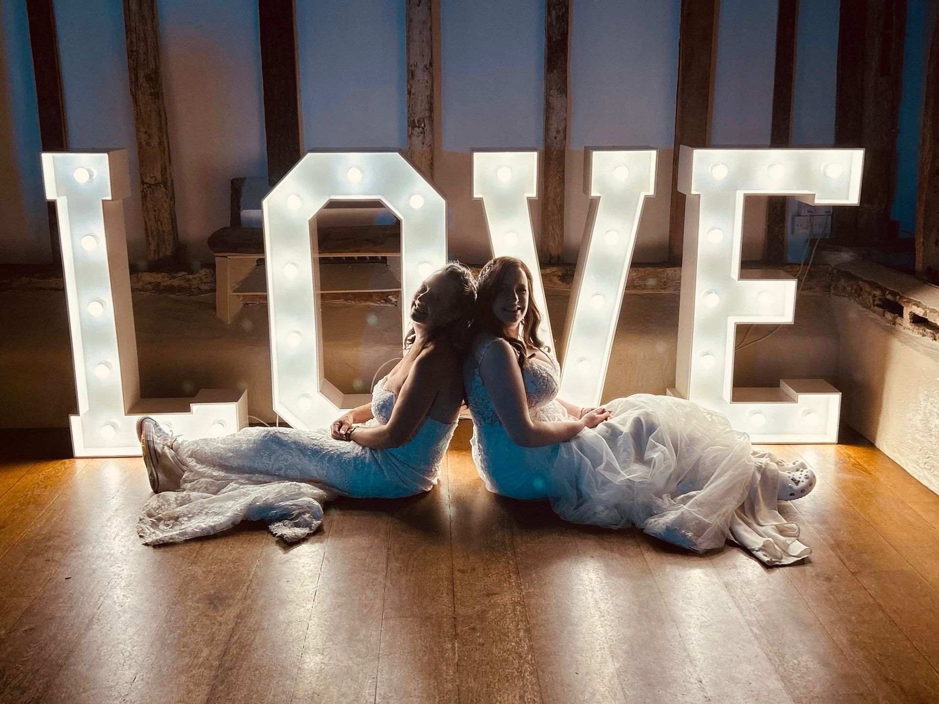 Two brides in white dresses on their wedding day, sat in front of our 4 foot tall LOVE letters that are turned on.