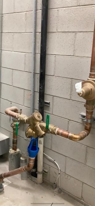 Plumbing pipes and valves against a cinder block wall, with a blue meter and copper piping in the foreground