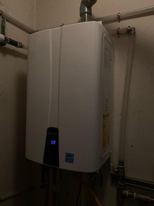 White wall-mounted water heater with a small blue display in a utility room, with pipes above and beside it