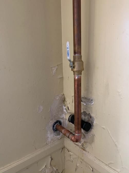 Exposed copper pipe and wall damage around plumbing hole in a beige interior corner