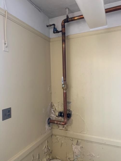 Exposed copper pipes running up a cream wall in a corner of a room beneath the ceiling.