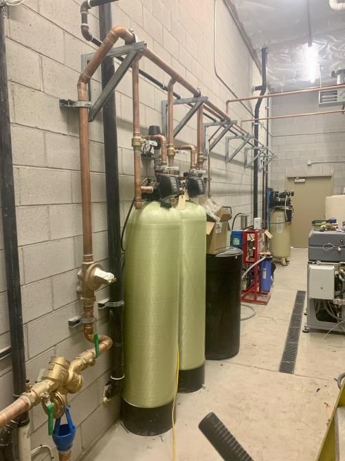 Industrial room with green and black tanks, pipes, valves, and equipment along a cinderblock wall.