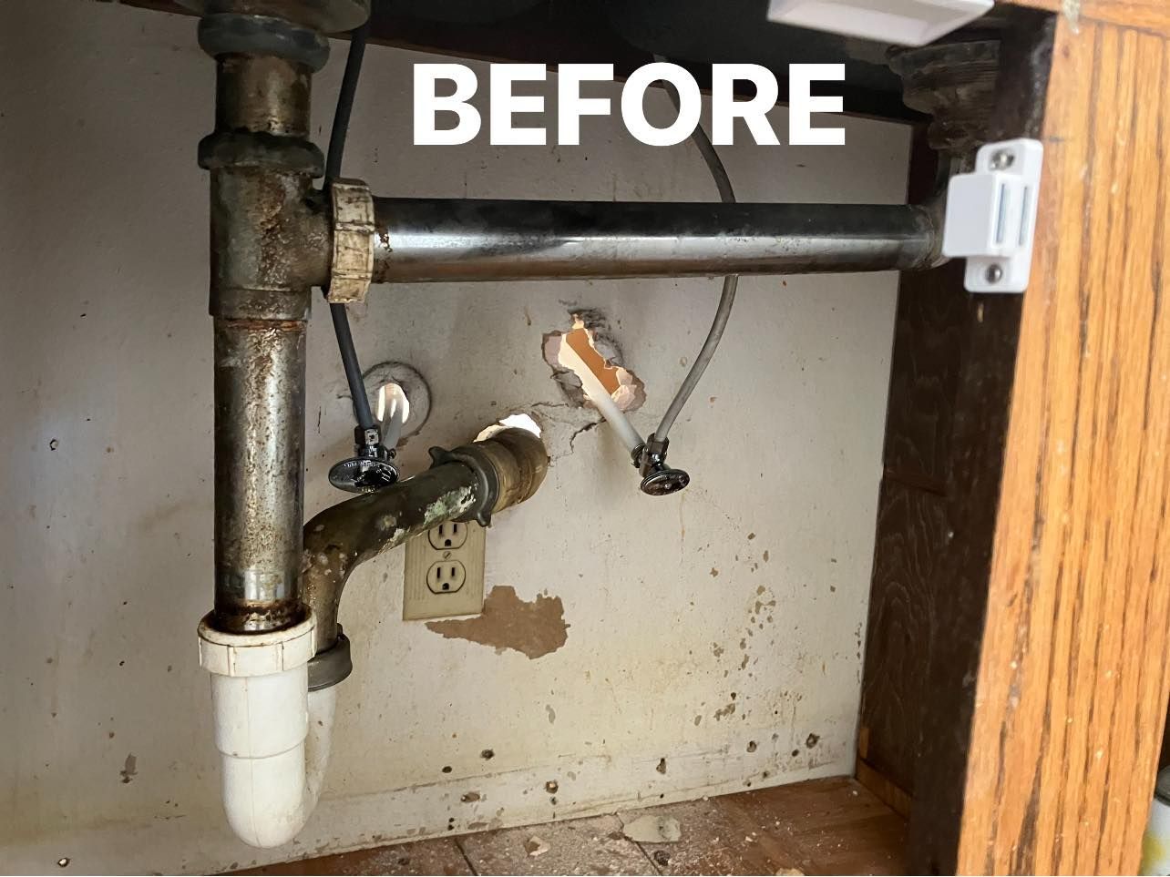 Under-sink plumbing before repair, with pipes, valves, and a water stain on the cabinet wall.