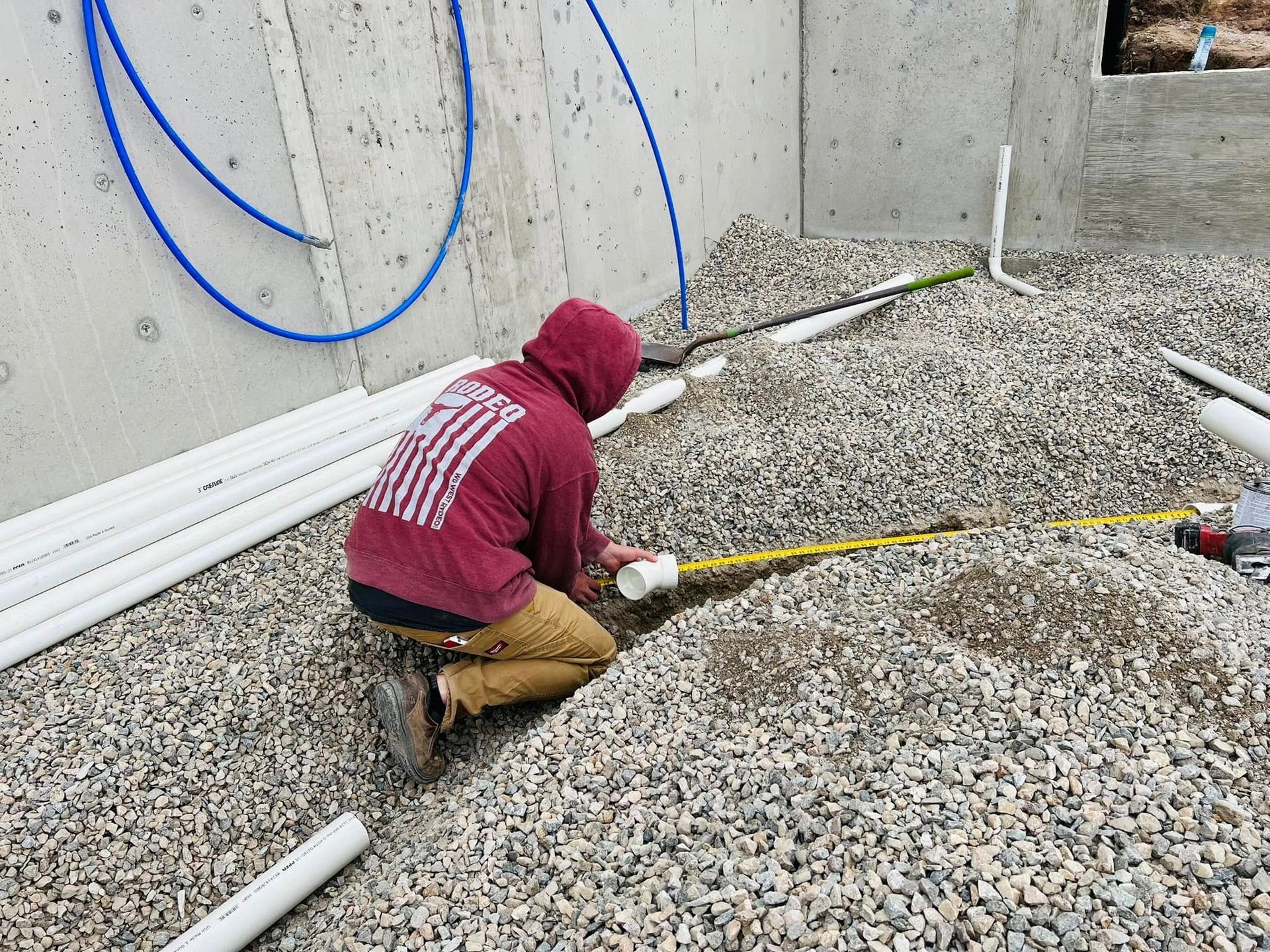 Worker in maroon hoodie crouches on gravel, handling a hose near exposed pipes at a construction site.