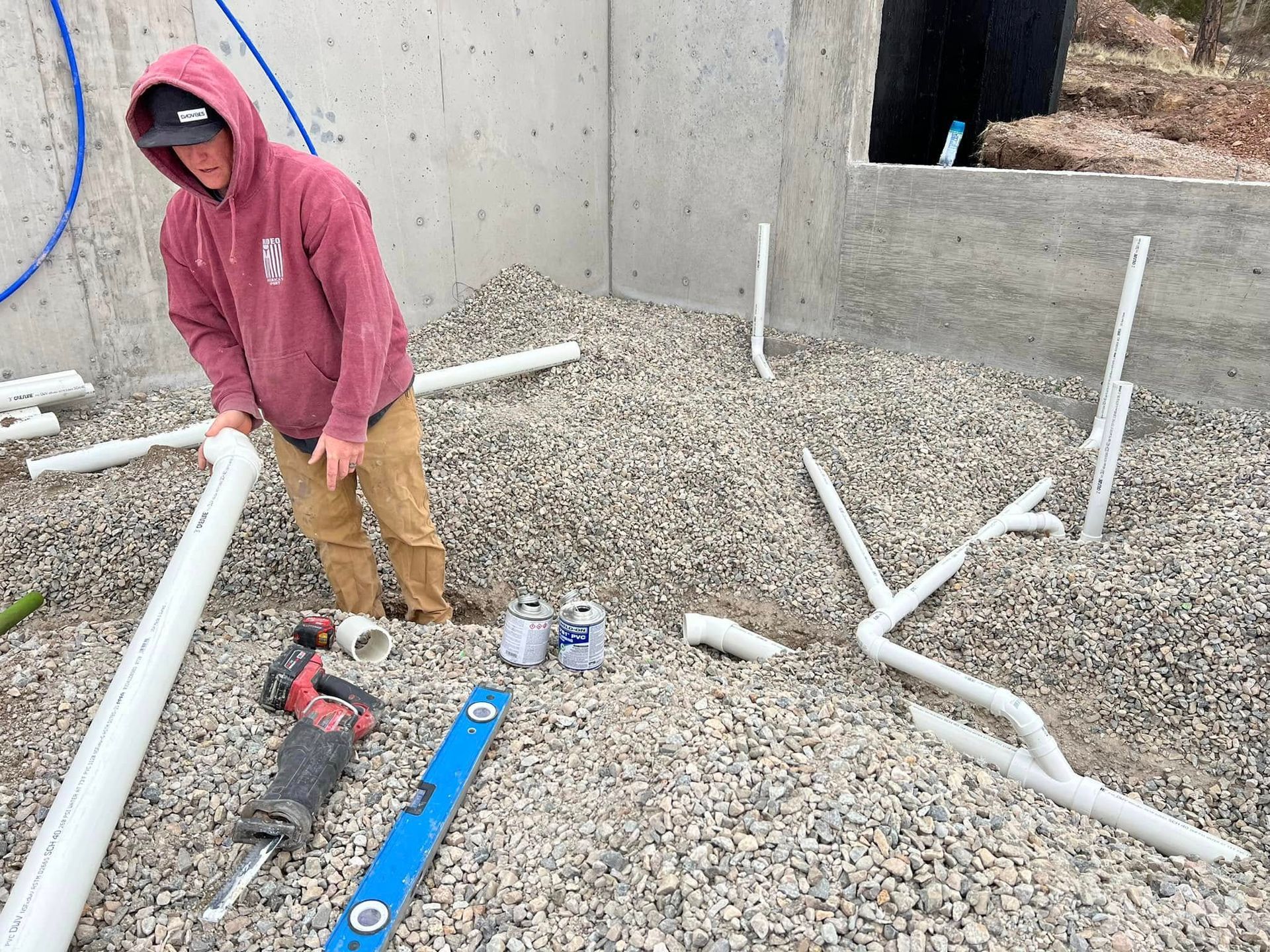 Person in a red hoodie standing among white PVC pipes and tools on a gravel construction site.