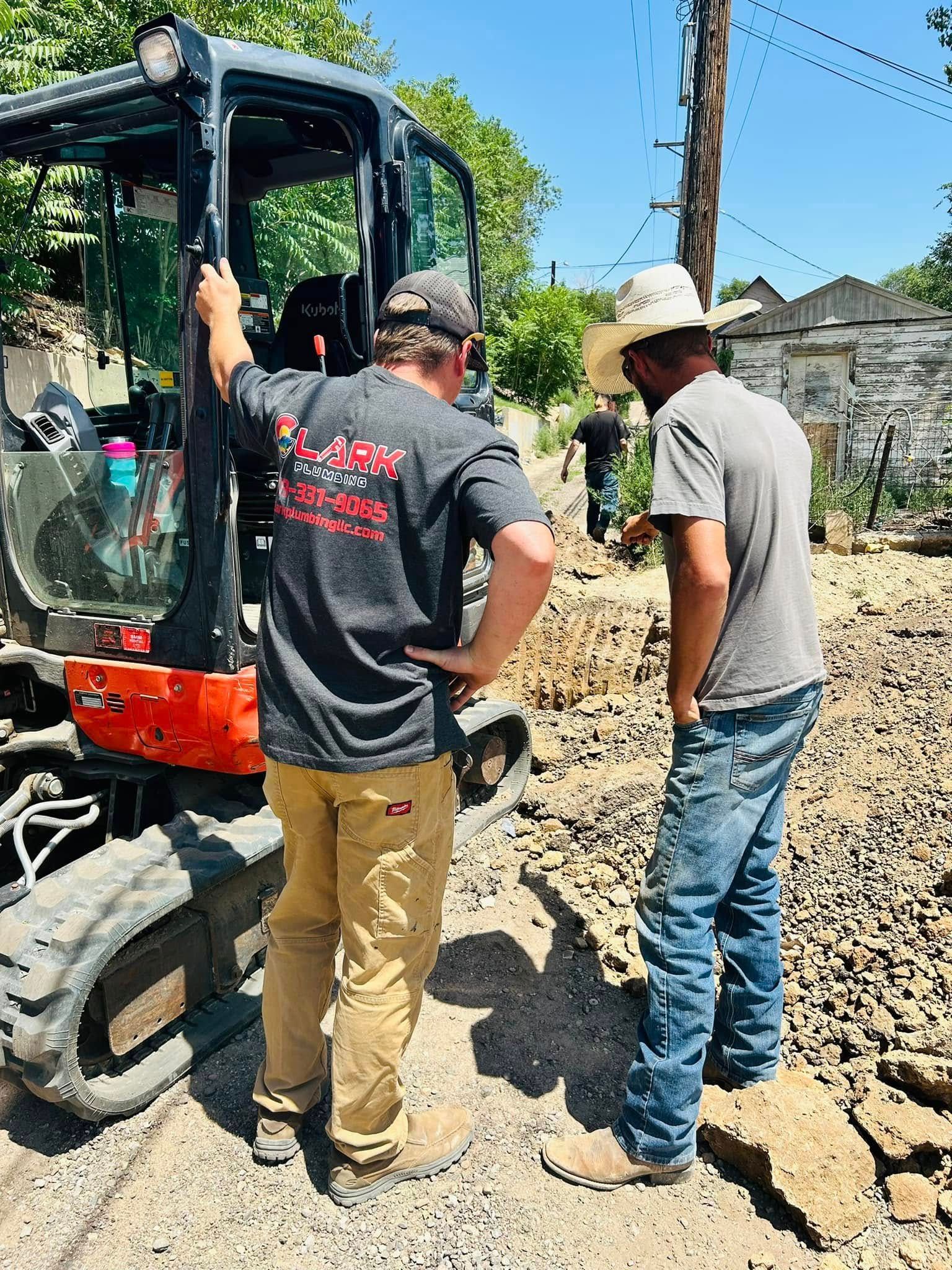 Two men inspect a compact excavator on a dirt site under a clear blue sky.