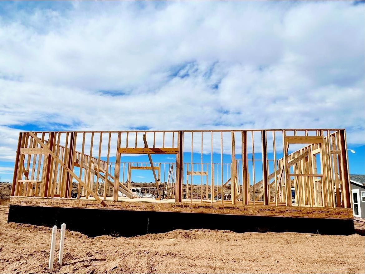 Wooden house frame under construction on a dirt lot beneath a blue, cloudy sky