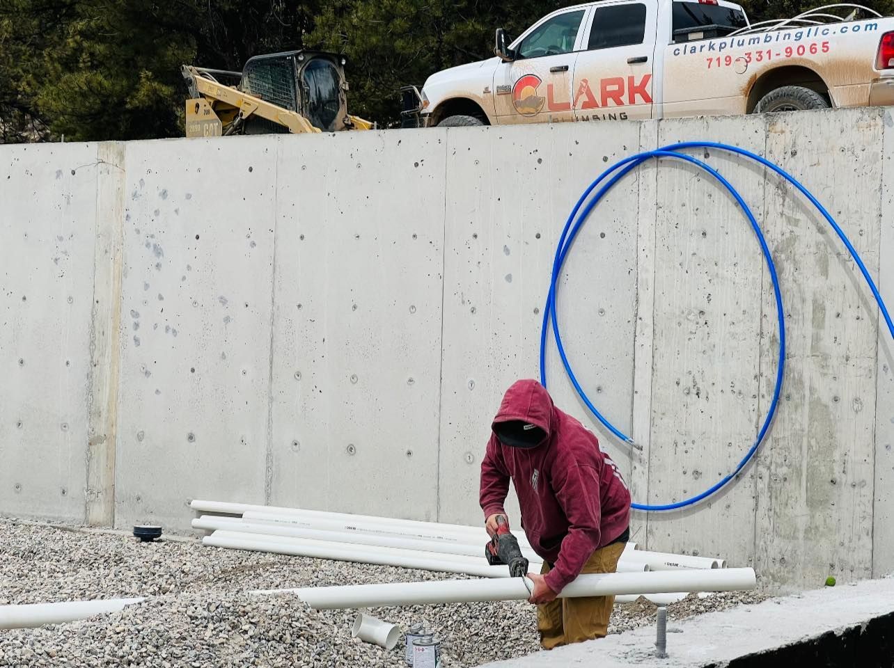 Worker in a red hoodie handling white pipes beside a concrete wall with a blue hose loop and parked truck above.