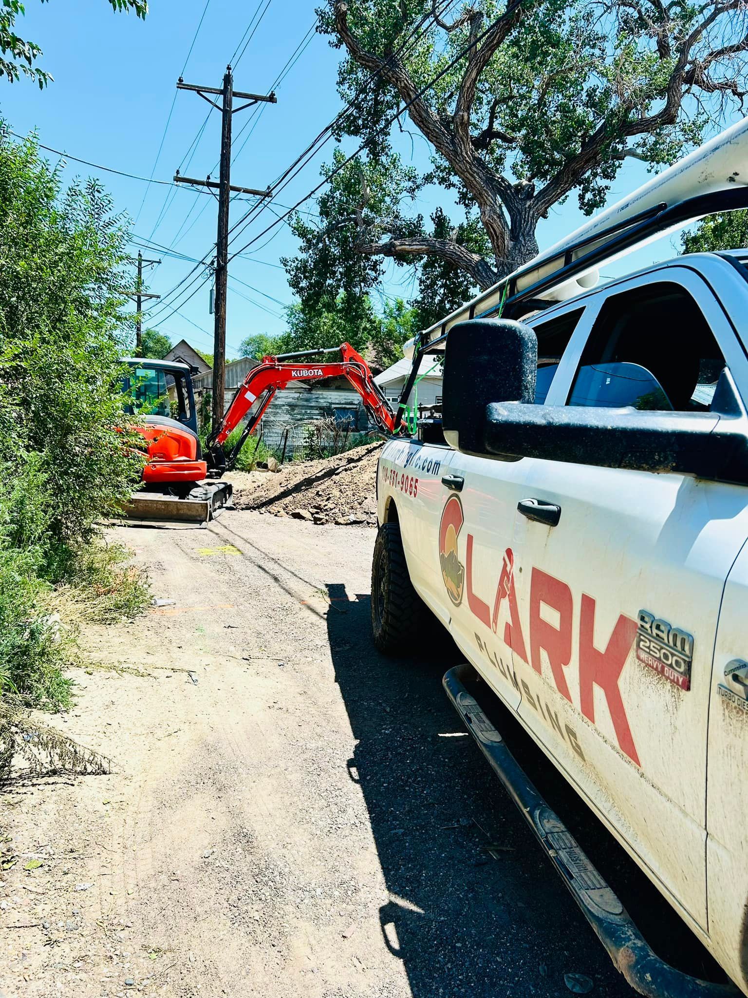 Roadwork with excavators and a white truck beside a gravel pile on a tree-lined street