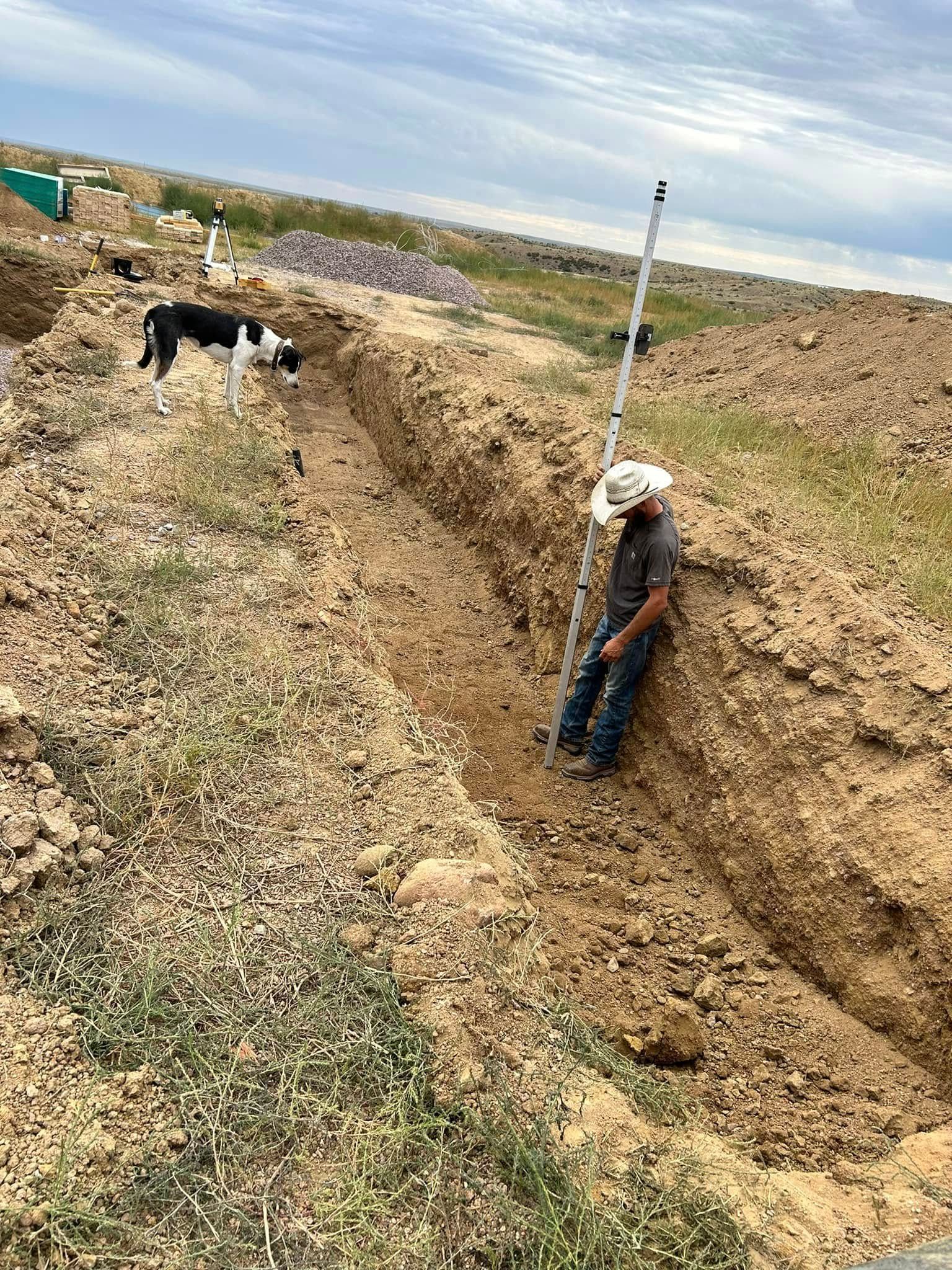 Person standing in a deep trench beside a dog in a dry field, with a marker pole nearby