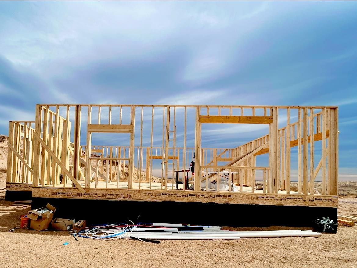 Wood-framed house under construction on a dirt lot beneath a blue sky