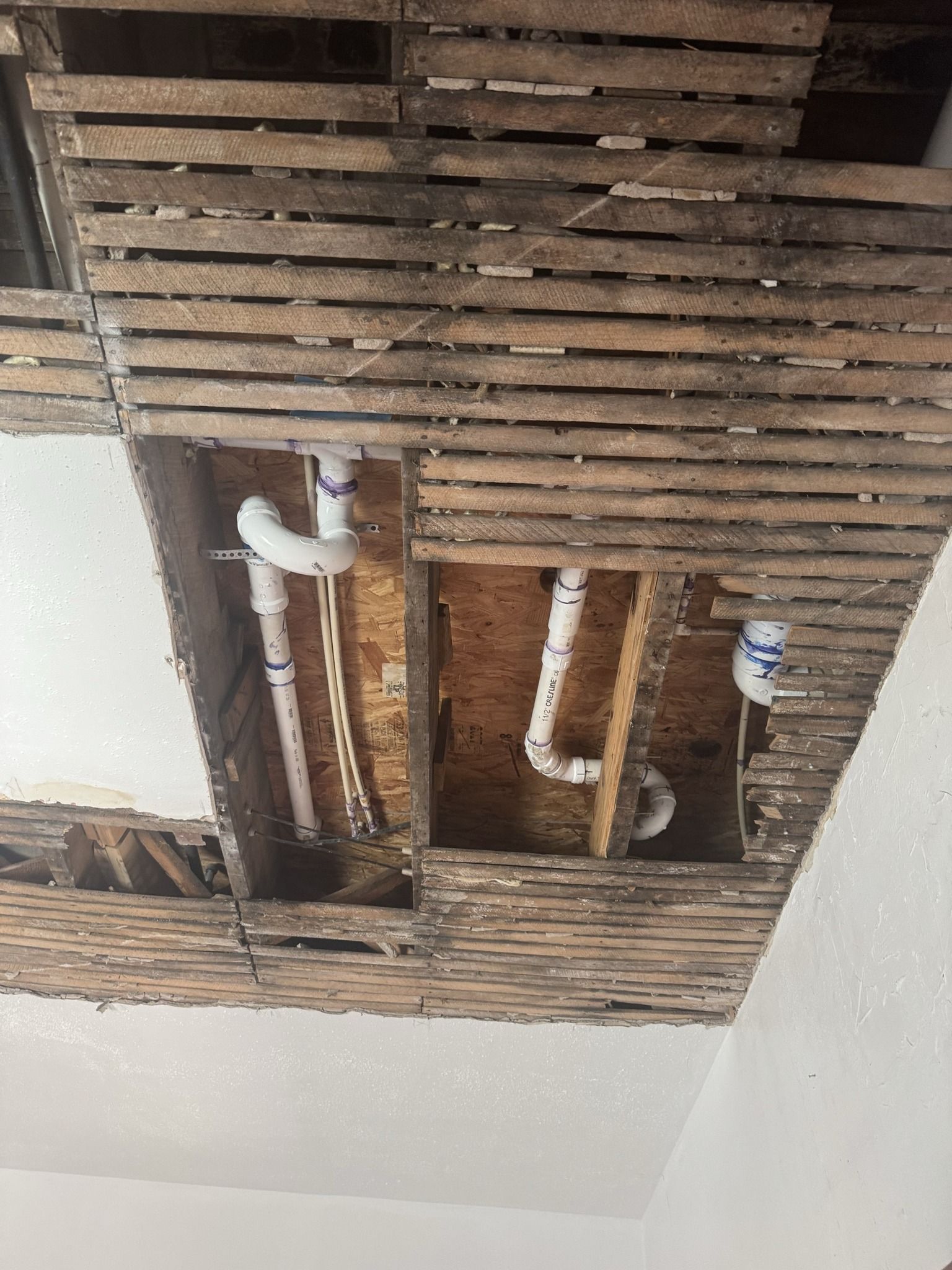 Damaged ceiling with exposed wooden joists, pipes, and a broken plaster opening