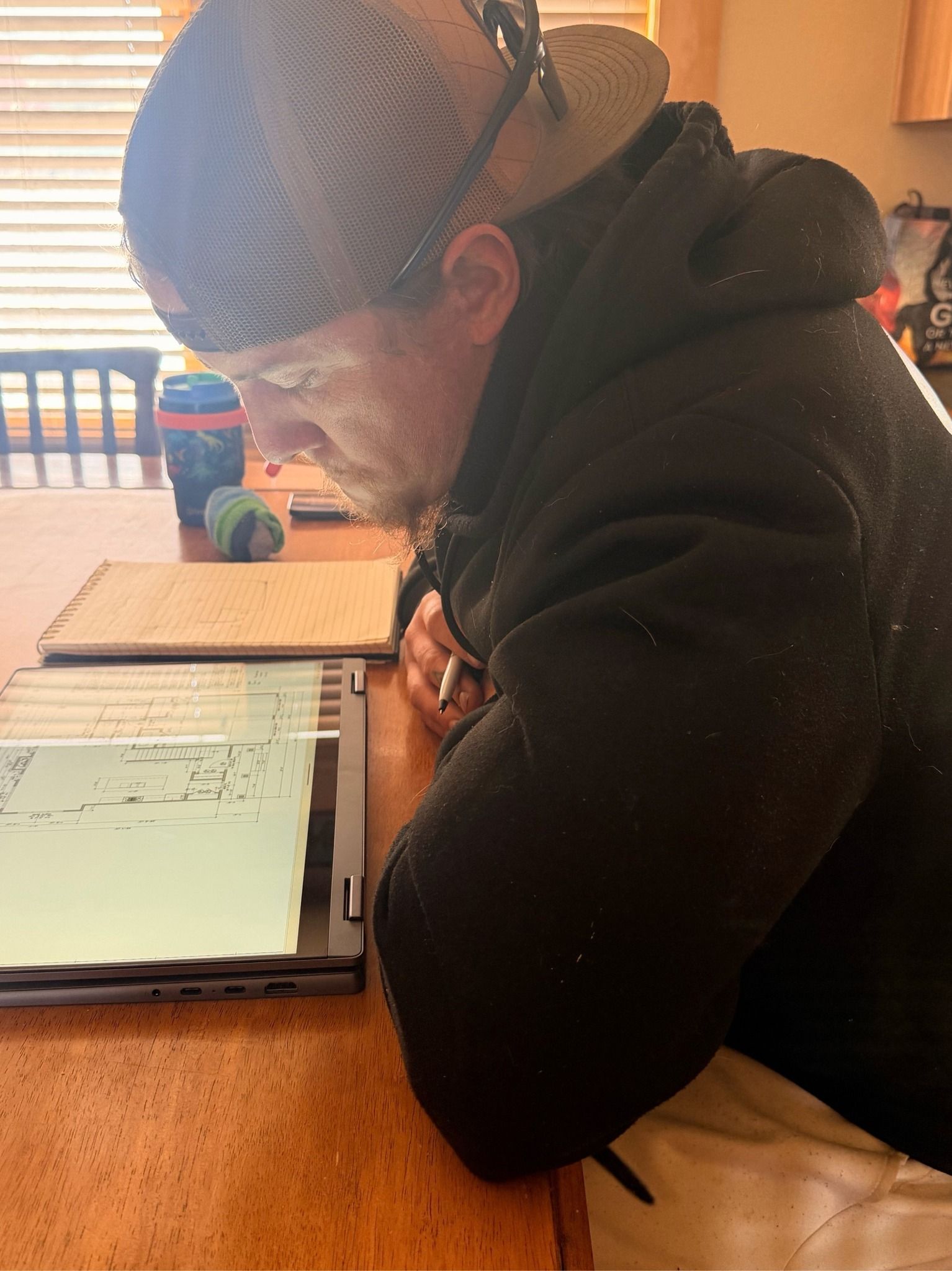 Person writing in a notebook at a wooden desk, wearing a black hoodie and blue head covering.