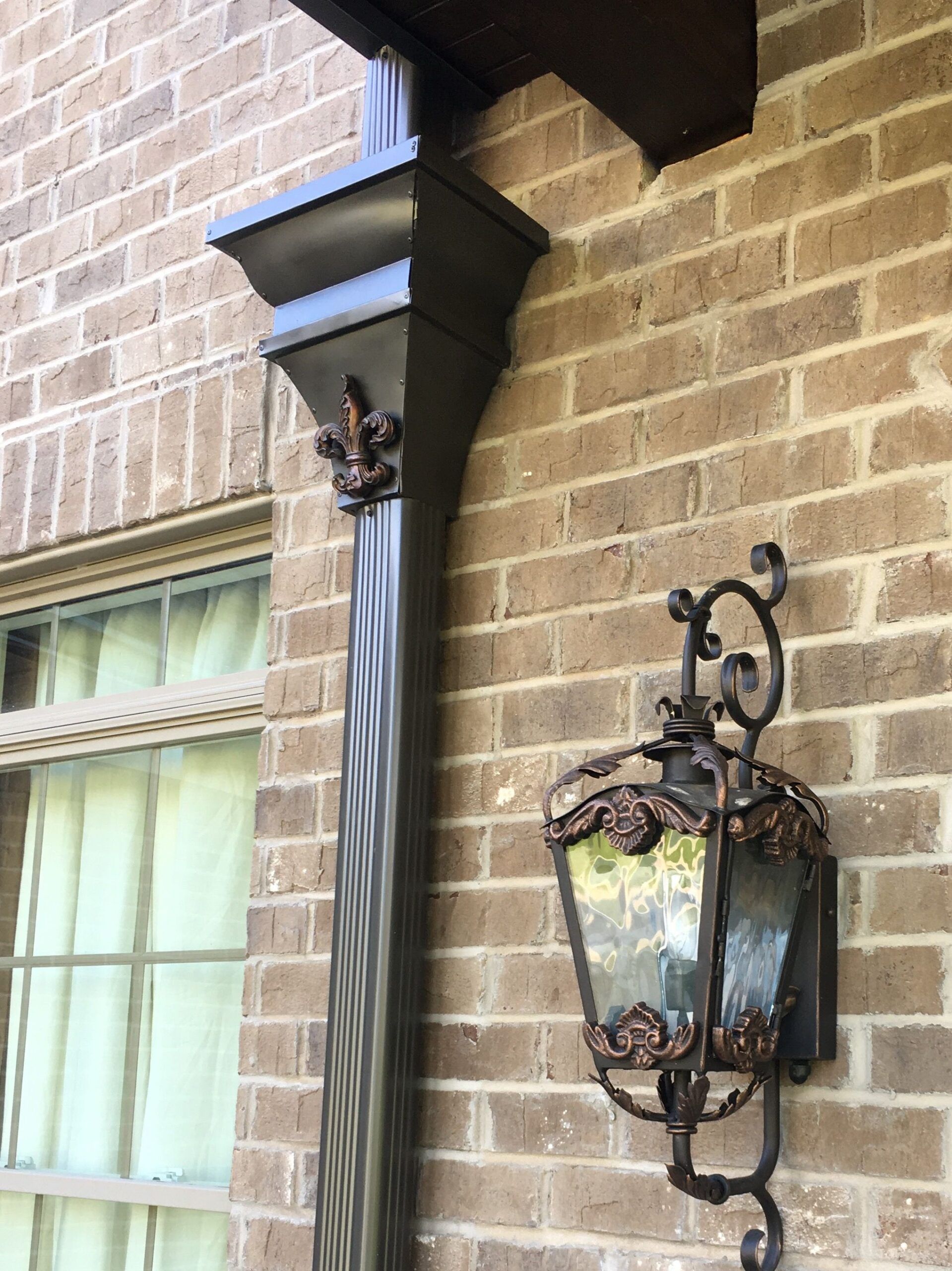 Brick house wall with an ornate black lantern and a dark column beside a window with curtains