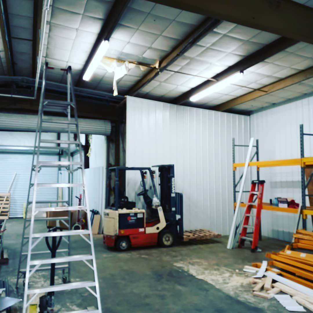 Warehouse interior with a forklift, ladders, metal shelving, and construction materials near a white wall
