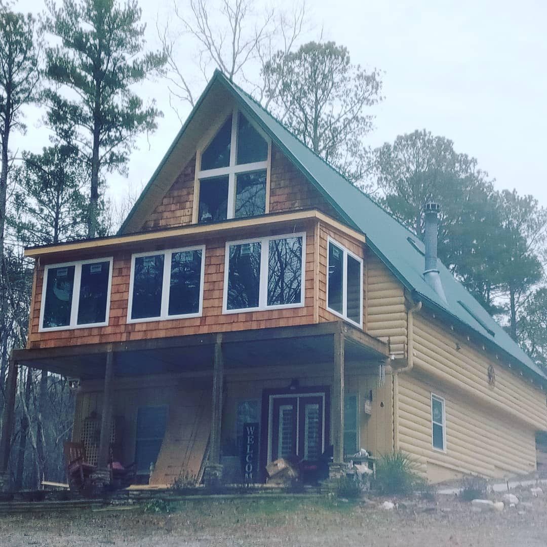 A rustic A-frame house with a porch, beige siding, and large windows in a wooded setting