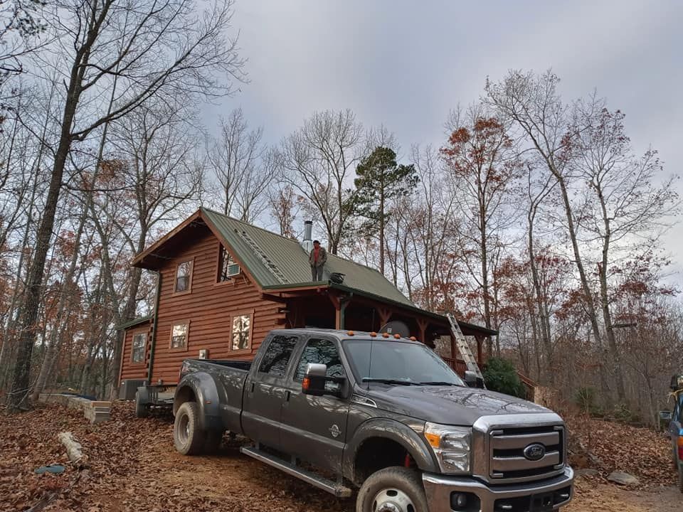 Brown cabin in a leafless forest with a black pickup truck parked in front.