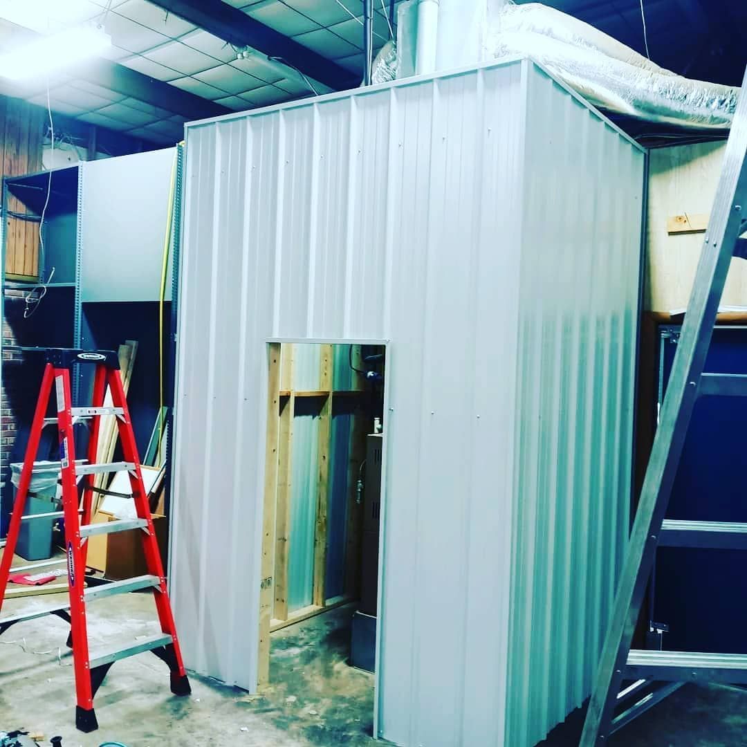 Small metal room under construction with open doorway, surrounded by ladders in an industrial space.