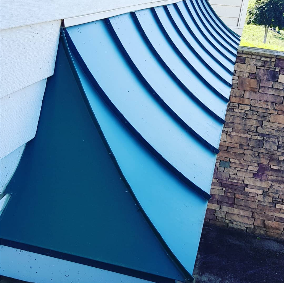 Curved blue metal roof panels beside a white wall and brick chimney