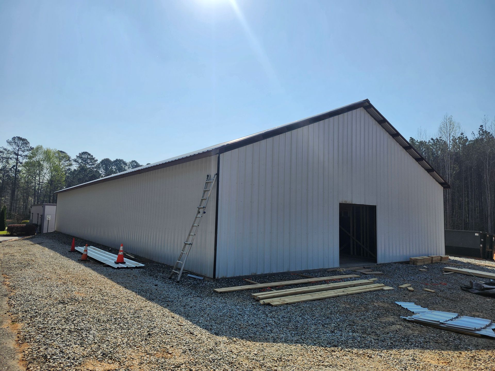 Large white metal building under construction on a gravel lot, with a ladder and traffic cones outside