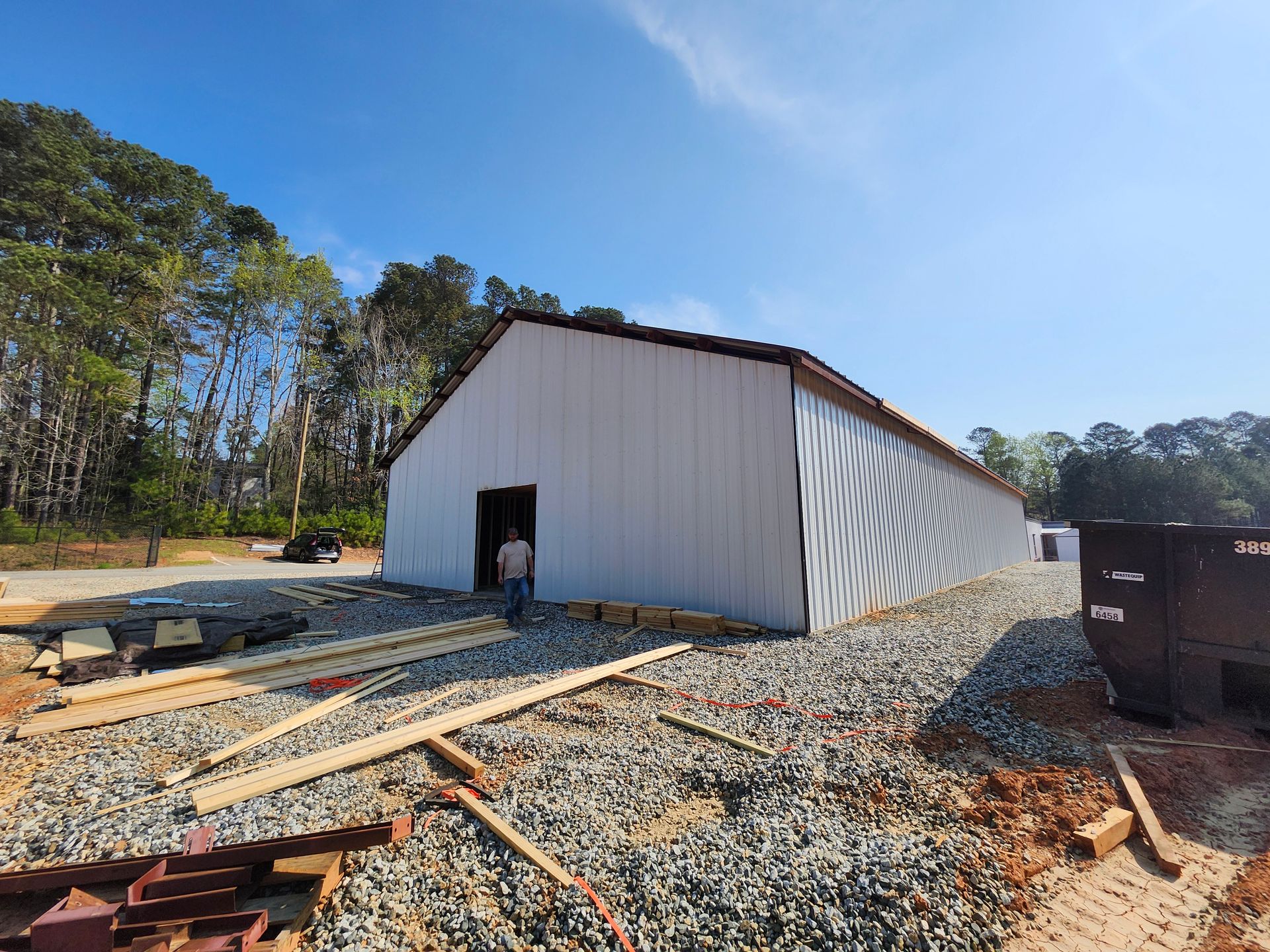 Partly built white metal shed on a gravel construction site with lumber and trees nearby
