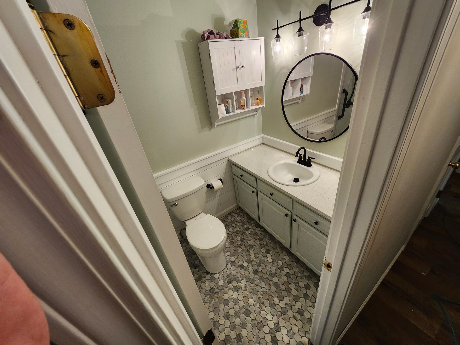 Small bathroom with white vanity, round mirror, toilet, and gray patterned floor tiles.