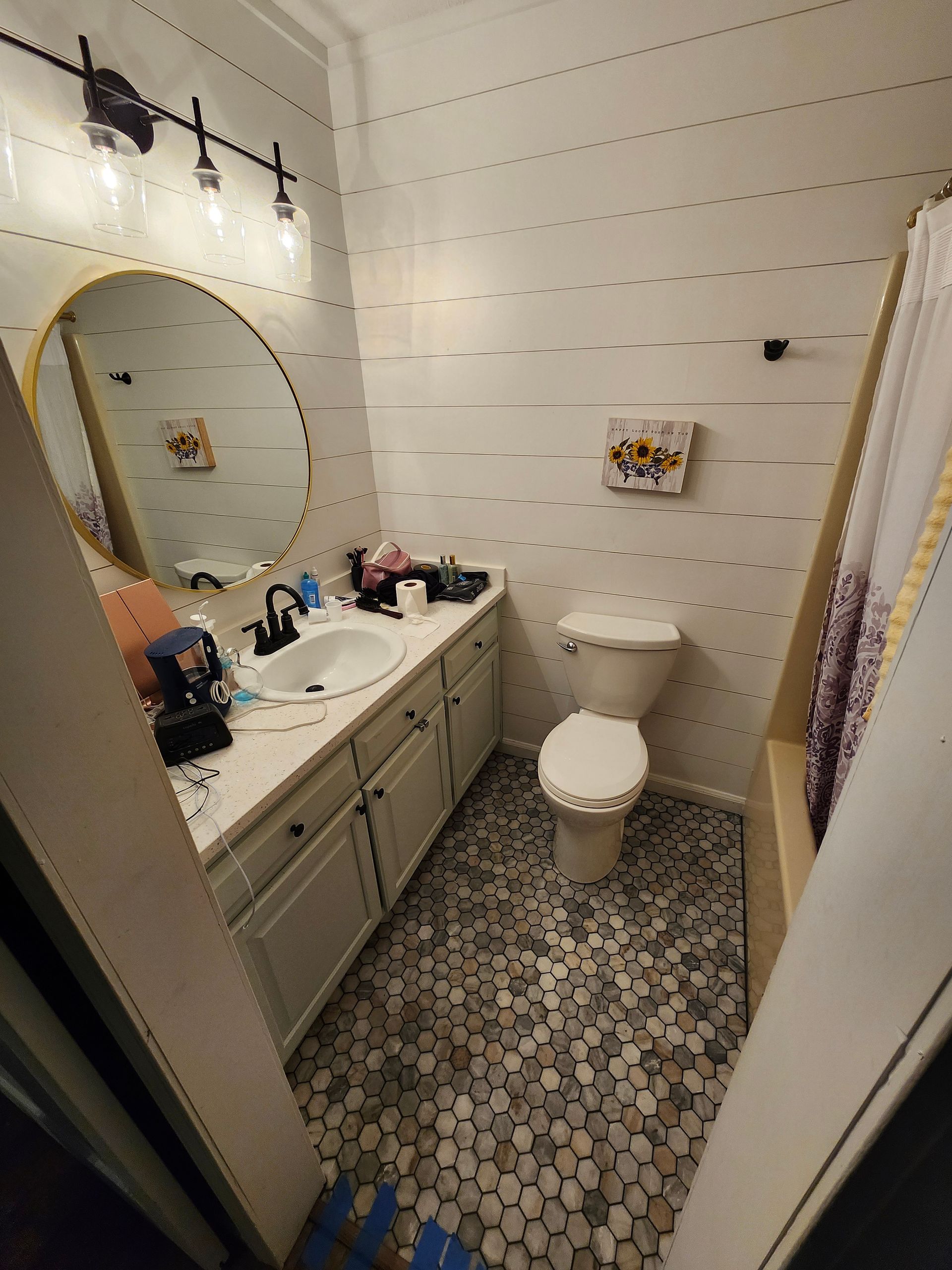 Small white bathroom with vanity, round mirror, toilet, patterned tile floor, and shower curtain.