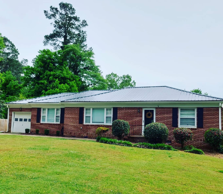Single-story brick house with a gray roof, green lawn, and trees in the background