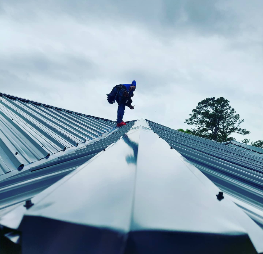 Person standing on a metal roof ridge under a cloudy sky, wearing a blue jacket and backpack