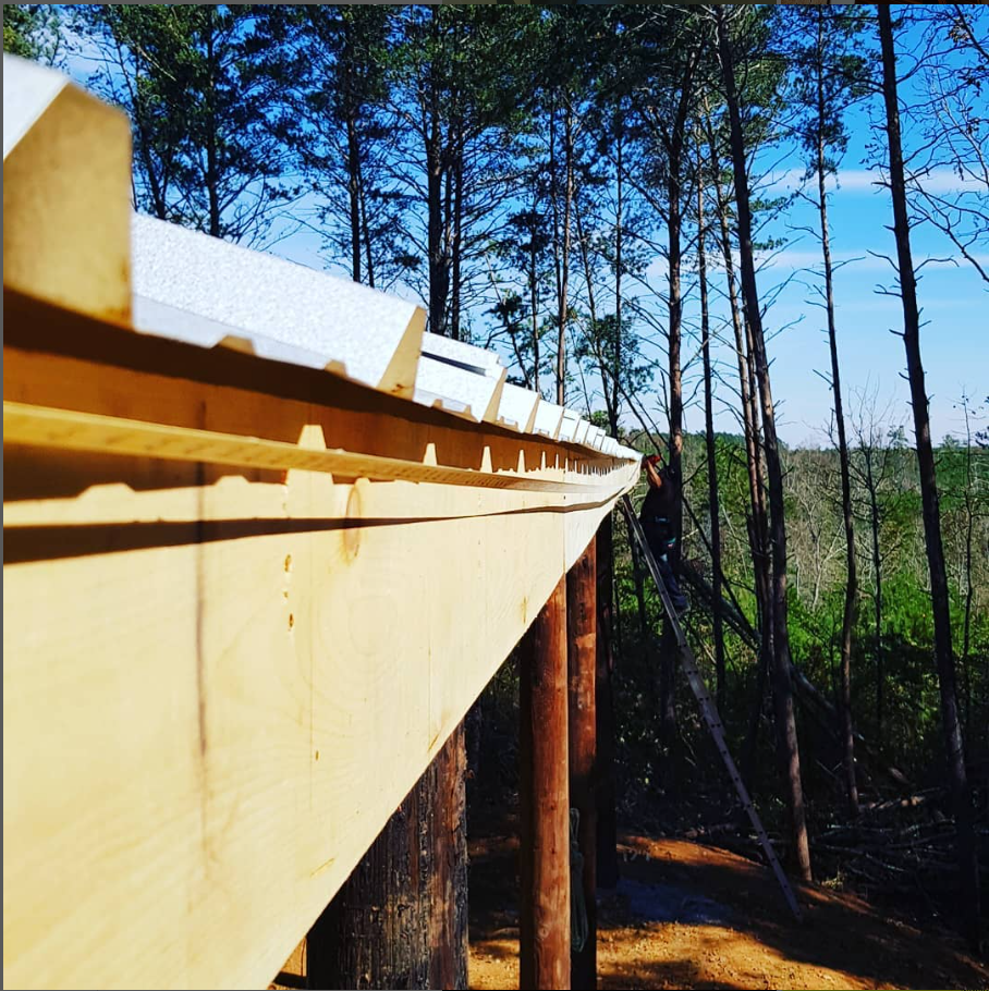 Wooden bridge under construction in a pine forest, seen from the side against a blue sky.