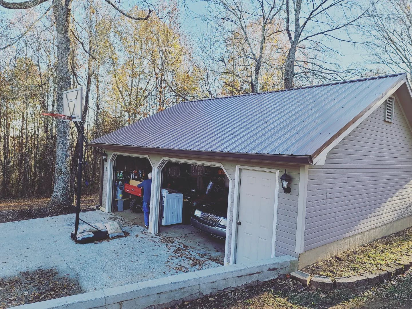 Detached gray garage with open doors, white shed, and gravel driveway beside leafless trees