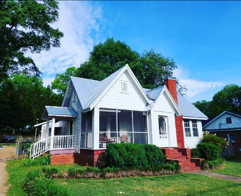 White cottage with a red brick foundation, screened porch, and green lawn surrounded by trees under a blue sky