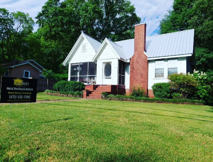 White house with green roof and brick chimney beside a lawn, with a black sign in front.