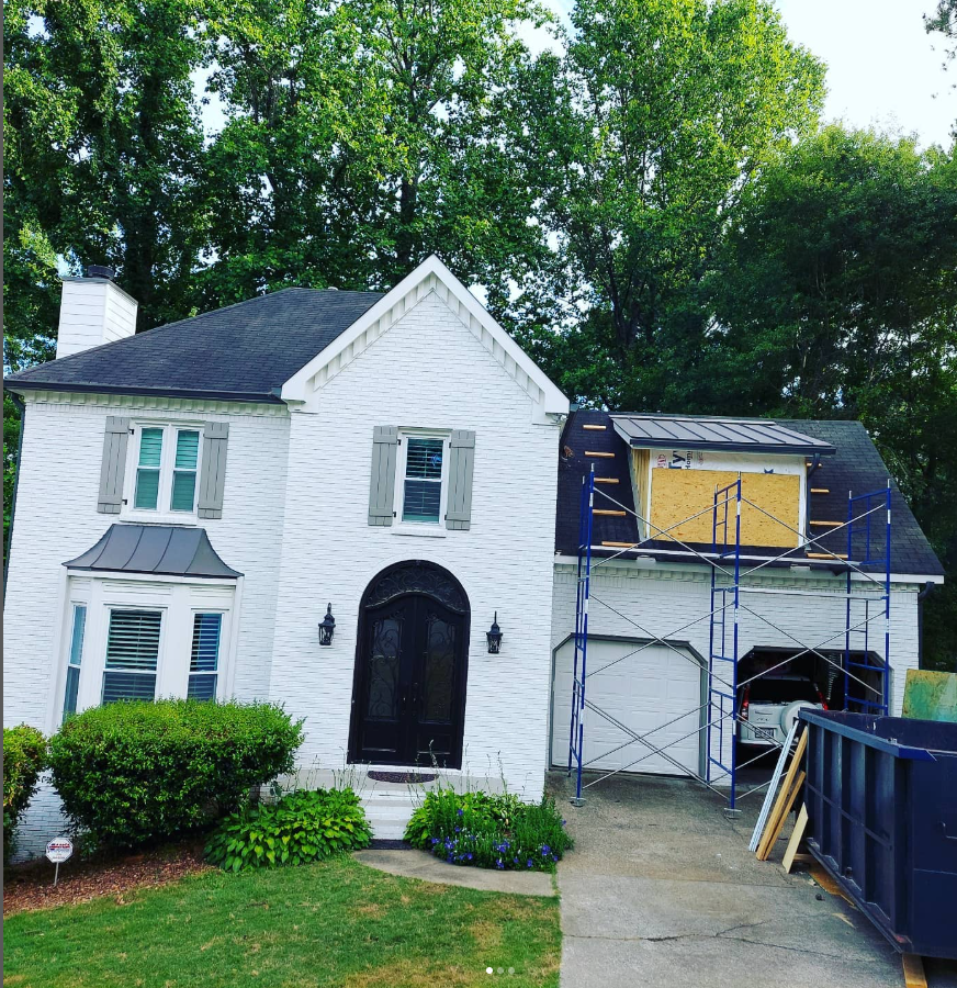 White brick house with black roof beside a scaffolded garage under renovation