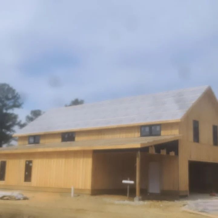 Partially built wooden house with a metal roof on a dirt lot under a cloudy sky