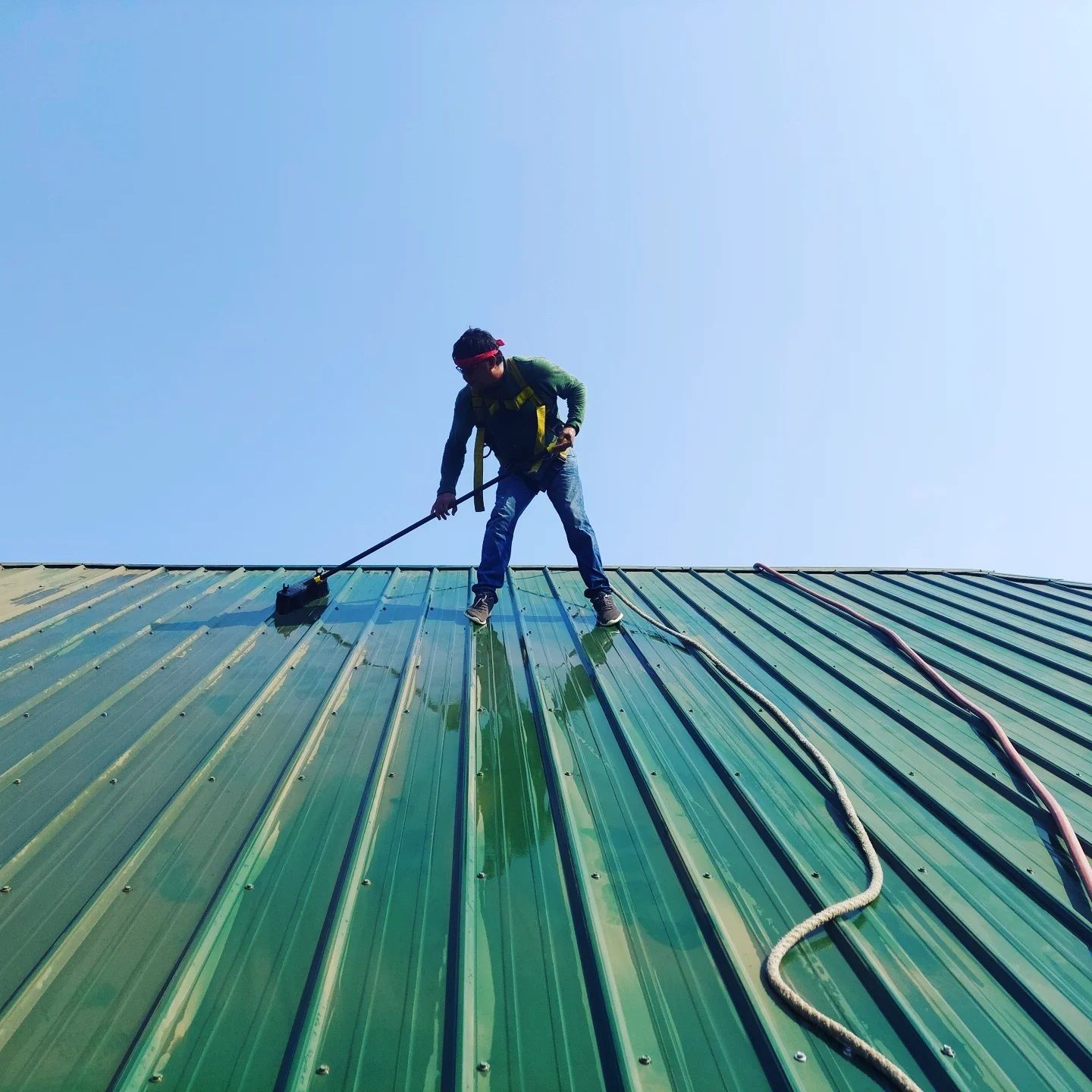 Worker cleaning a green metal roof with a hose under a clear blue sky
