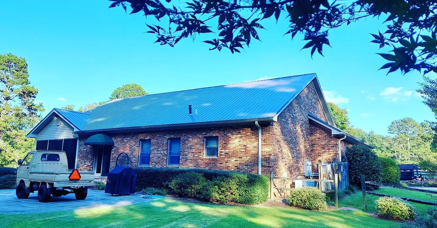 Stone lodge with blue roof in a grassy yard, with a parked vehicle and trees in bright daylight